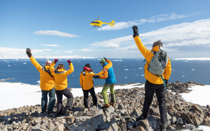 Group of guests in yellow coats stand atop rocky summit & wave to yellow helicopter on a sunny day in Antarctica.
