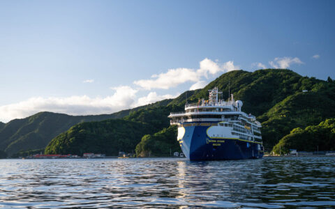 Modern blue & white expedition ship sits in calm sea beside rows of green mountains under a nearly clear sky in Japan.