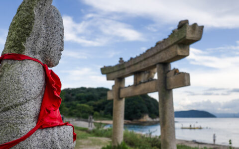 Stone Buddha statue with red shirt tied over its front sits near a stone torii gate beside a white-sand beach in Japan.