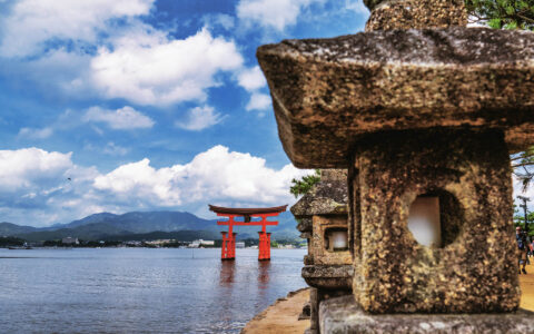 Red wooden torii gate sits in calm sea backed by green mountains & framed by weathered stone mini pagoda statues in Japan.