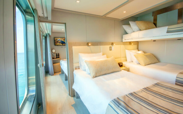 Triple cabin with 3 twin-sized beds with white linens connected to an adjoining room aboard small ship Origin Theory.