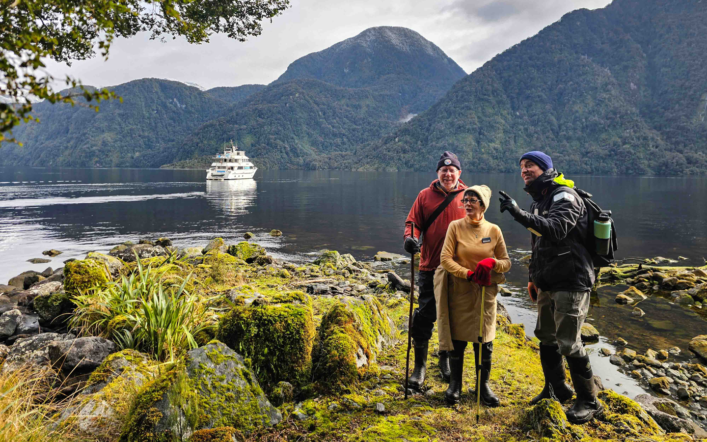 Woman in yellow & man in red stand by guide on mossy ground with small white ship nearby on a Doubtful Sound overnight cruise.