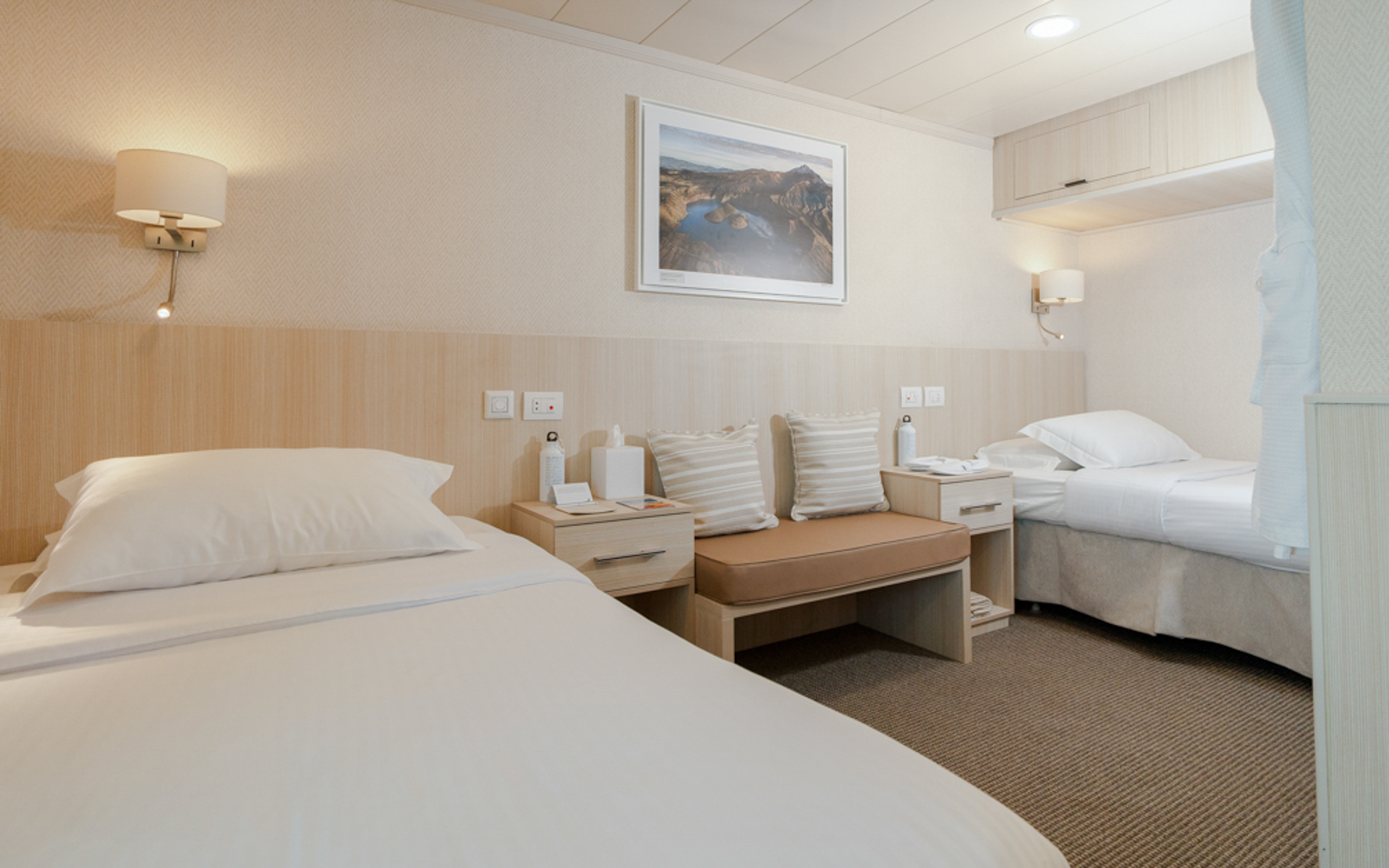 Small ship Isabela II stateroom double bed with white linens, bench seat, overhead storage, nightstands and reading lights.