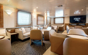 Salon bar aboard Small ship Isabella II with grey couches, brown leather chairs, tables and large viewing windows.