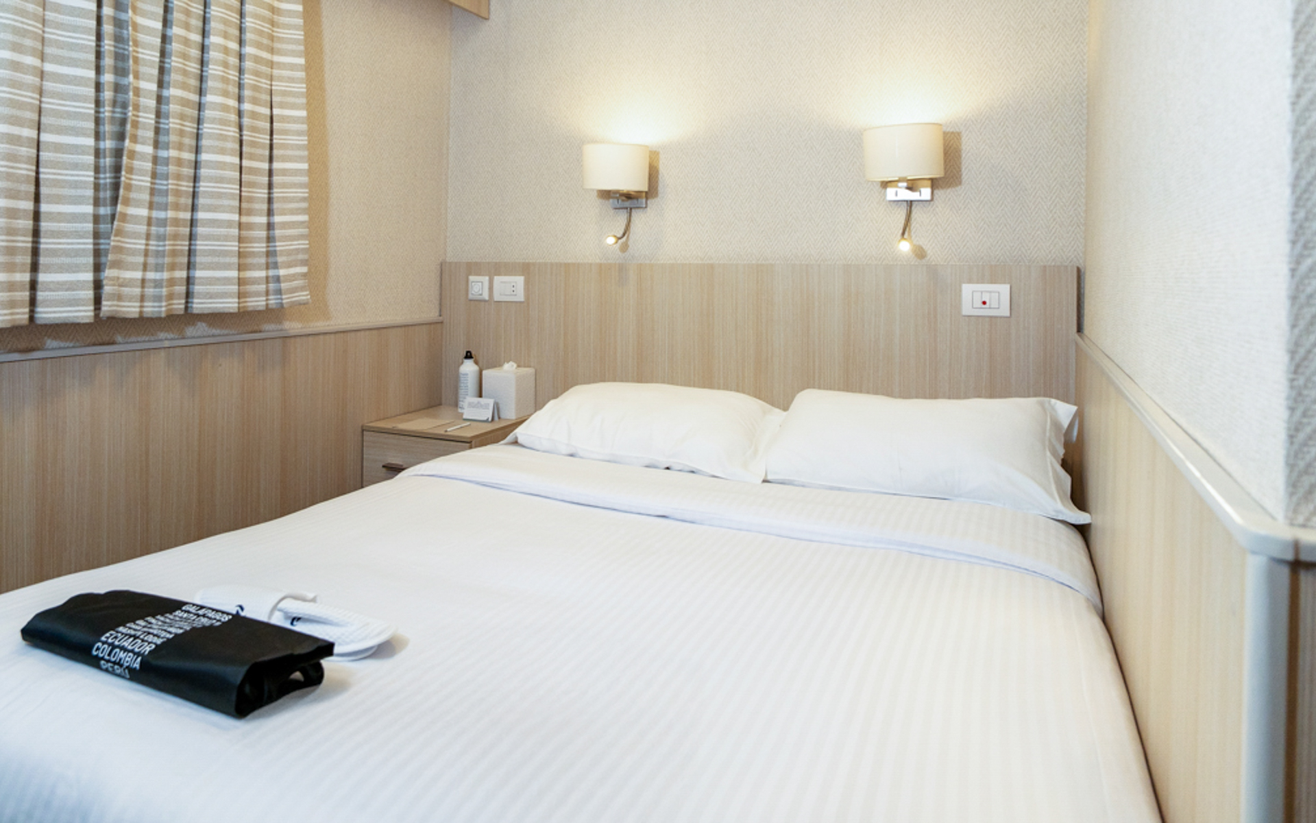 Small ship Isabela II stateroom double bed with white linens, window, nightstand &two reading lights. Slippers & laundry bag on bed