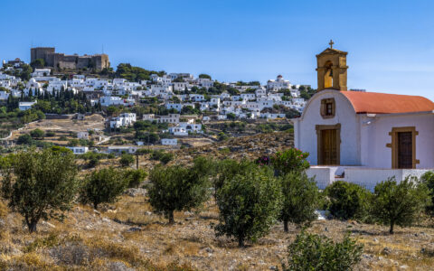 Small whitewashed church with red roof & small belltower beside olive grove & hillside town of white buildings in Greece.