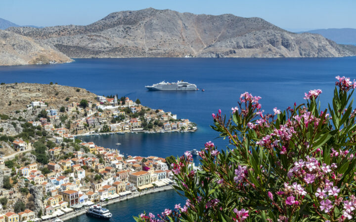 Aerial view of pink flowers overlooking seaside Greek island town with small gray expedition ship sitting offshore in the sun.
