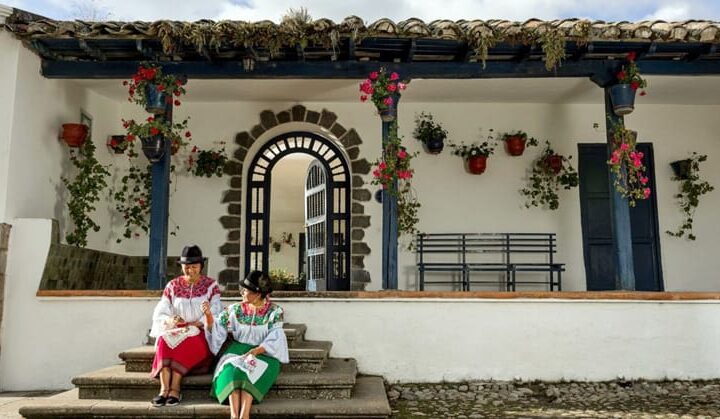 2 Ecuadorian women in traditional clothing sit on the entry steps to Hacienda Zuleta, a white building with blue wood accents & potted flowers