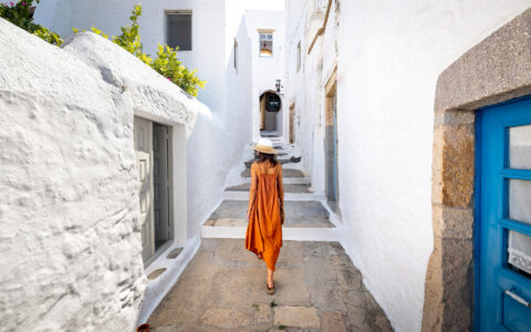Woman in orange dress, white & tan hat walking through narrow path between white washed buildings during Greek Island small ship cruise