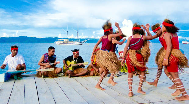 Local children dancing in their traditional ceremonial attire on a dock with a small ship cruise in the background in Indonesia.
