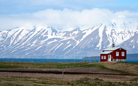 Small red house sits on stilts overlooking blue water with snowcapped peaks in the background, seen on an Iceland cruise.