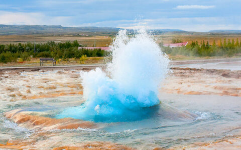 Geothermal pool explodes with icy-blue scalding water amidst tangerine sand & sediment in front of vast fields in an expansive valley in Iceland.
