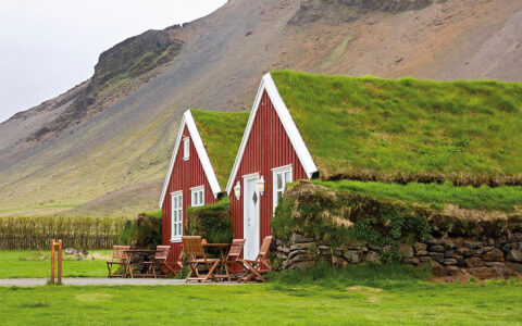 2 red buildings with their roofs covered in grass in a vineyard at the base of a sweeping mountain. 2 outdoor tables and 6 chairs sit by building