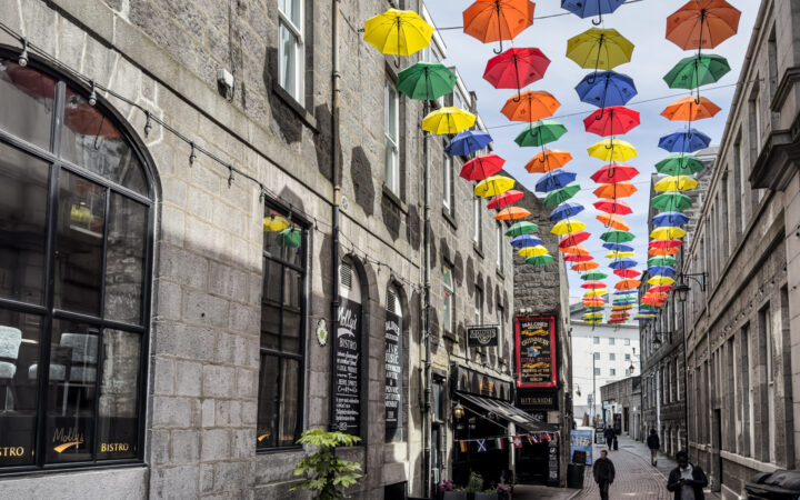Individuals walk a brick road between tall stone buildings & under rows of hanging colorful umbrellas in Aberdeen, Scotland.