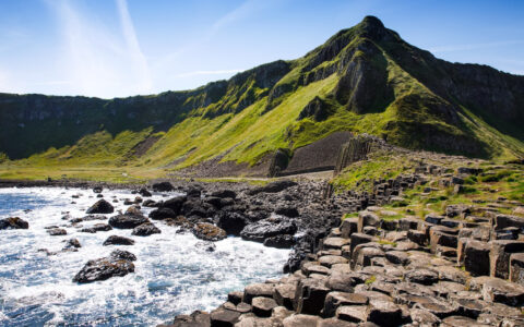 Northern Ireland's Giant's Causeway trail with brown basalt columns by green mountains & black volcanic peaks by the sea.