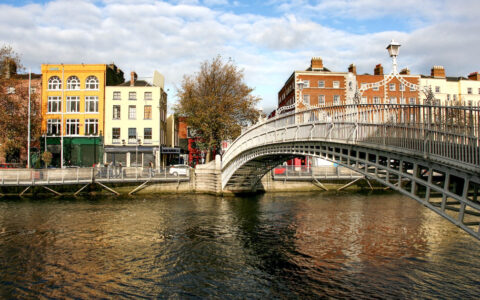 Steel Ha'Penny Bridge in Dublin, Ireland extends over a calm canal backed by 4-story buildings of brick on a sunny day.