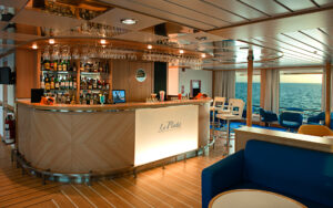 Bar aboard La Pinta yacht. Floor-to-ceiling windows along walls, room filled with low dining tables. A few bar stools at the bar.