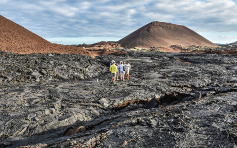 La Pinta Galapagos Cruise travelers walk over hardened black lava field near brown mountains on a cloudy day.