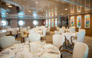 Dining area showing 8 tables set for a meal next to large port holes aboard La Pinta small cruise. Reflective ceiling and warm lighting.