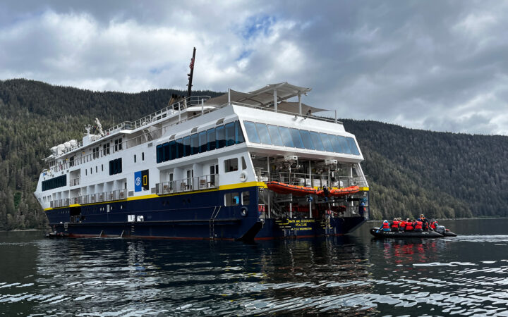 Port side & rear of Nat Geo Venture small ship as a Zodiac pulls up in calm water on a cloudy day in Alaska.