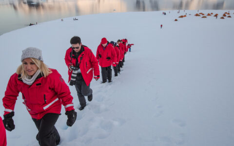 Group of hikers in red jackets walks up a snowfield away from tents at dusk on the Quest For The Antarctic Circle cruise.