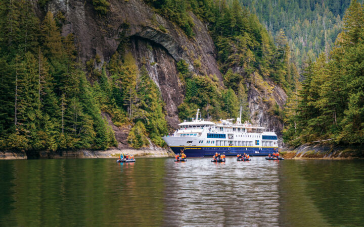 Small ship with Blue hull & white upper decks sits in glassy water in an inlet in Alaska as Zodiacs with travelers motor by.