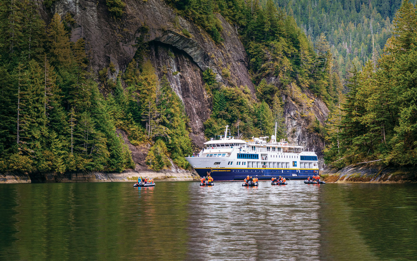 Small ship with Blue hull & white upper decks sits in glassy water in an inlet in Alaska as Zodiacs with travelers motor by.