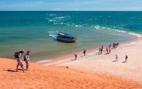 Travelers walk down a red sand hill towards people on the beach as a small aluminum boat pulls into shore in west Australia.