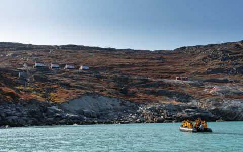 Zodiac boat with guests heads to autumnal arctic tundra via turquoise water on a sunny day during West Greenland small ship cruise