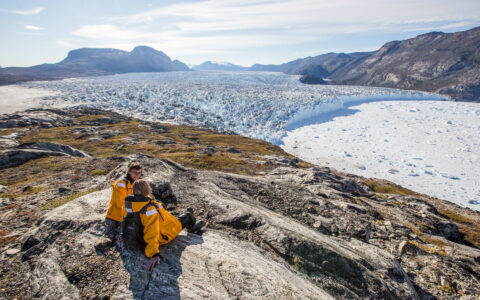 2 female Arctic travelers in yellow parkas sit atop a mountain of rock & tundra overlooking a large glacier during West Greenland small ship cruise