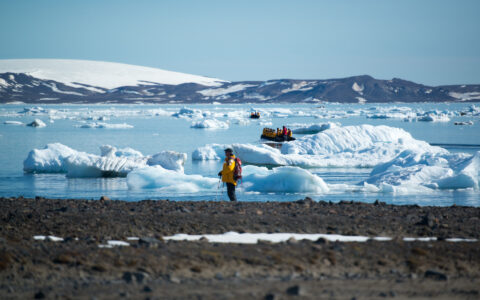 Arctic traveler in yellow parka stands on rocky shore as Zodiacs with others sail around an icy bay during West Greenland small ship cruise