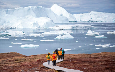 Arctic travelers walk wooden boardwalk over tundra toward iceberg-choked bay of calm water during West Greenland small ship cruise