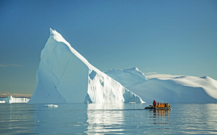 Zodiac with travelers passing massive pointed iceberg rising out of calm water on a sunny day during West Greenland small ship cruise