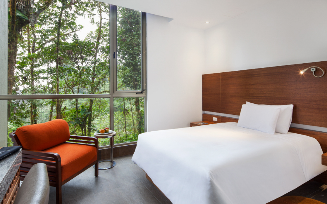 King-sized bed in single room next to orange armchair and and a private patio overlooking the jungle at the Mashpi Lodge in Ecuador.
