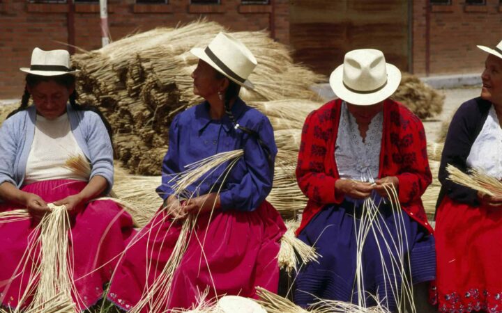 ecuadorian women sit in a line weaving straw with a bushel behind them