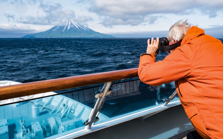 Man in orange jacket stands on deck of small ship & photographs volcano across dark blue sea on an Alaska to Japan cruise.