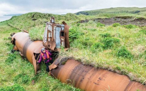 Woman in purple coat & orange life jacket stands in doorway of rusted submarine surrounded by overgrown green grass,