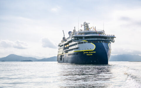 Modern small ship National Geographic Resolution with sharp bow, dark blue hull & white upper decks sits in calm sea.