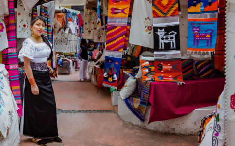 Woman shopkeeper stands in open-air market amongst brightly colored woven textiles, seen on the Andes Highland Haciendas tour.