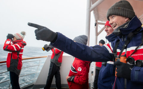 Explorers dressed for the cold weather in gloves and hats look out off the stern of a small cruise ship