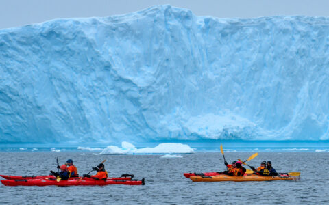 cold weather Kayakers navigate icy waters in front of massive iceberg thats light blue in color