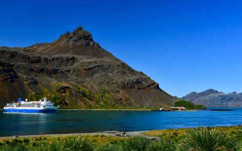 Small cruise ship sits in the bay of blue waters surrounded by steep mountains on a blue sky day