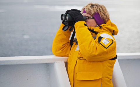 Woman in yellow parka stands on deck of ship looking through binoculars on an Arctic Saga: Spitsbergen, Faroes & Jan Mayen cruise.
