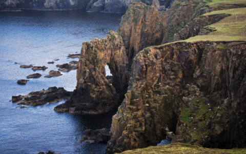 Aerial view of cliffs leading into the sea with one forming an arch & green grass above, seen on an Arctic Saga cruise.