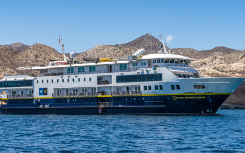 National Geographic Venture small ship with blue hull & white upper decks sits offshore of arid beige mountains in Baja.