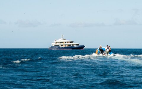 Small ship with red & blue hull & white upper decks waits for travelers approaching on a Zodiac on a Horizon Galapagos cruise.
