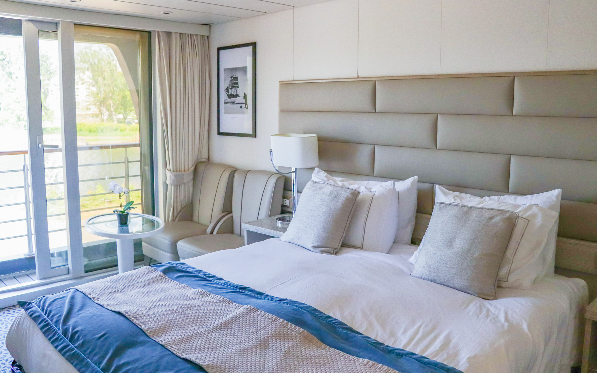 Balcony Stateroom aboard Discoverer: Polar Latitudes on small ship showing a large bed fitted with white & tan linens next to chairs, desk & sliding glass balcony doors
