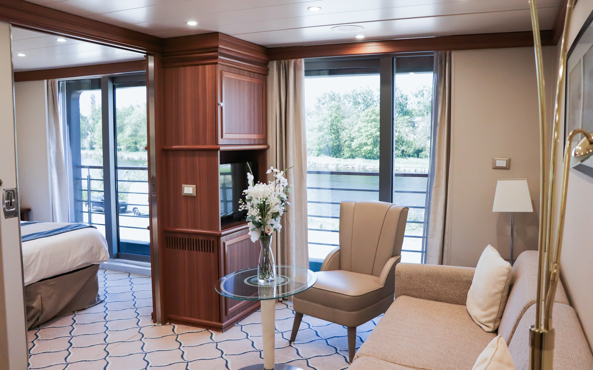 Spacious Discovery Suite aboard Discoverer: Polar Latitudes with bed to the left & couch with chair & table to the right separated by TV cabinetry