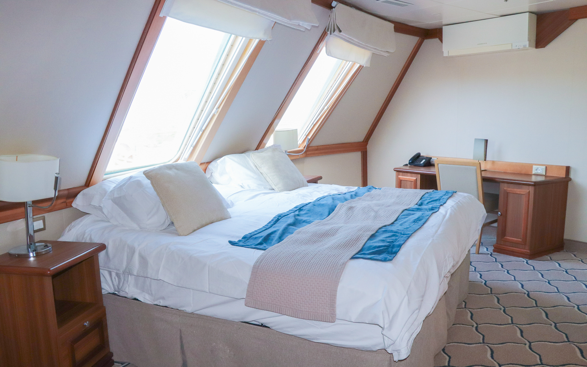 Grand Suite aboard Discoverer: Polar Latitudes complimented by wood accents and two overhanging windows shows bed fitted with white & tan linens aboard small ship.