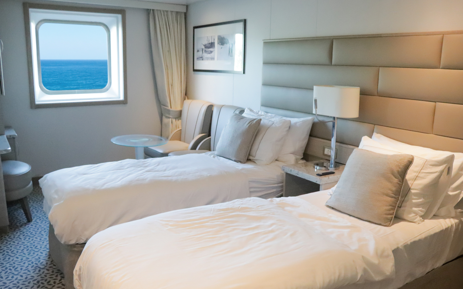 Ocean Stateroom on small ship showing a large bed fitted with white & tan linens next to couch, desk, vanity, stool & large window